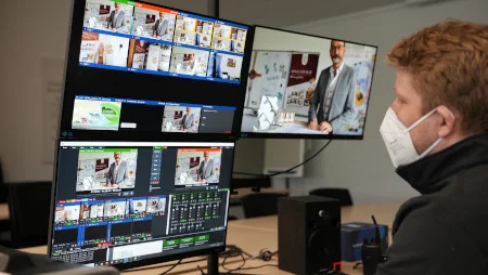 Mann mit Mundschutz sitzt an einem Tisch vor diversen Bildschirmen und Übertragungstechnik. Auf einem der Bildschirme ist GEPA-Geschäftsführer Peter Schaumberger zu sehen.