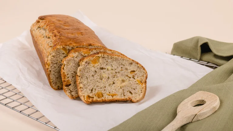 Auf wei&szlig;em Papier auf einem Rost liegt ein Bananenbrot, von dem zwei Scheiben schon abgeschnitten sind. Daneben liegt dekorativ ein olivgr&uuml;nes Tuch mit einem Holzl&ouml;ffel darauf.