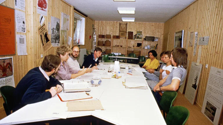 Sichtlich altes Bild, auf dem acht junge Menschen an einem Konferenztisch sitzen und diskutieren. Vor ihnen liegen aufgeschlagene Aktenordner und Papiere.