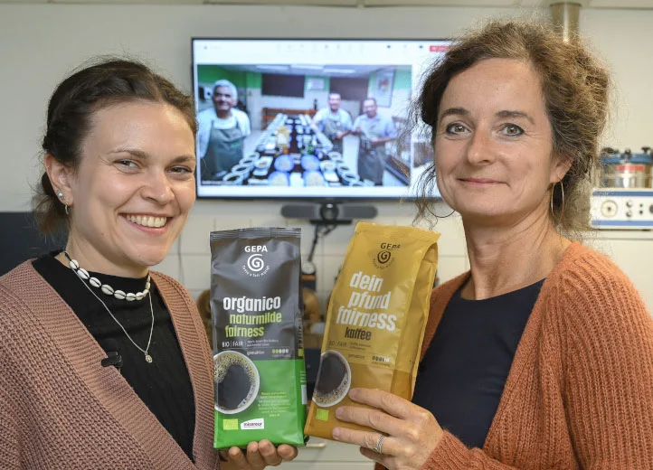 Zwei Frauen in einem Kaffeelabor lächeln und halten je eine Packung GEPA-Kaffee in die Kamera. Hinter ihnen auf einem Bildschirm sind per Videocall drei Männer zugeschaltet, die sich auch in einem Kaffeelabor befinden.