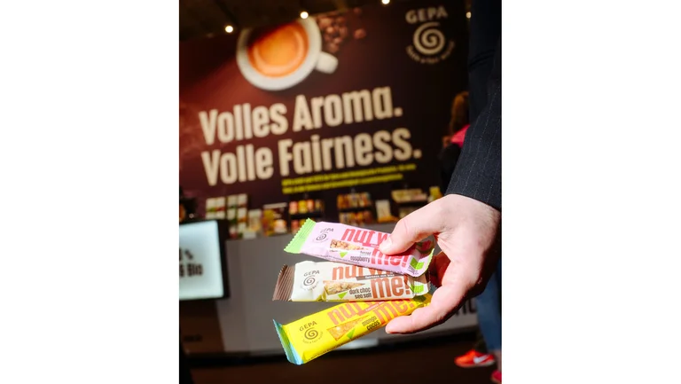 Hand h&auml;lt drei verschiedene GEPA-Nussriegel mit der Aufschrift "nut with me!". Im Hintergrund an einer Messewand steht "Volles Aroma. Volle Fairness."