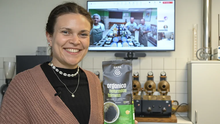 Eine Frau in einem Kaffeelabor lächelt und hält eine Packung GEPA-Kaffee "organico" in die Kamera. Hinter ihr auf einem Bildschirm sind per Videocall drei Männer zugeschaltet, die sich auch in einem Kaffeelabor befinden.
