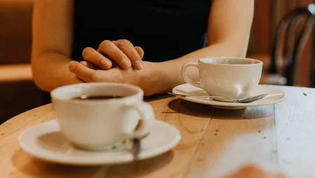Zwei Tassen Kaffee stehen auf einem runden Holtzisch. Man sieht die Arme einer Frau, die ihre Arme auf dem Tisch abst&uuml;tzt.