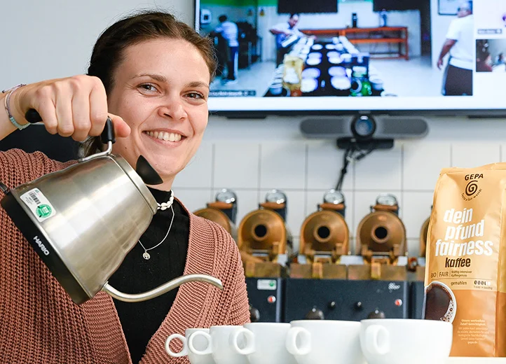 Junge Frau in einem Kaffeelabor h&auml;lt eine silberne Kanne &uuml;ber wei&szlig;e Tassen, als w&uuml;rde sie Kaffee aufgie&szlig;en, und l&auml;chelt in die Kamera. Am Rand steht eine Packung GEPA-Kaffee "dein pfund fairness".