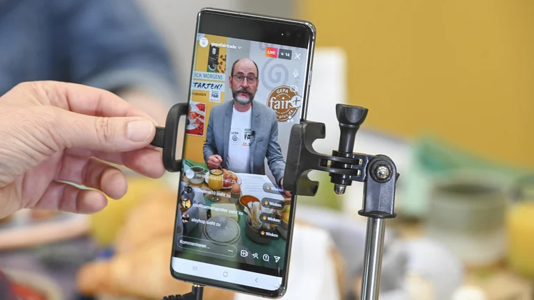 Nahaufnahme eines Smartphones, das von einer Hand gehalten wird. Im Display sieht man GEPA-Gesch&auml;ftsf&uuml;hrer Peter Schaumberger, der an einem Fr&uuml;hst&uuml;ckstisch sitzt und in die Handykamera spricht.