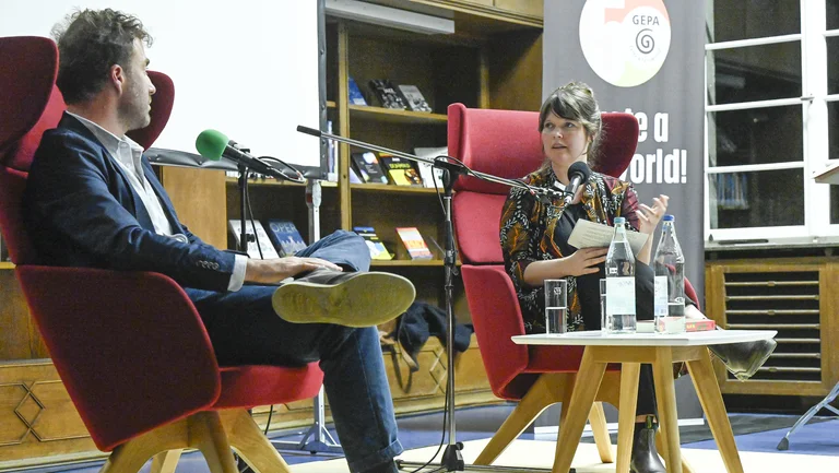 Mann und Frau sitzen in Sesseln vor einem Bücherregal und führen einen Dialog. Sie sprechen in Mikrofone.