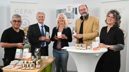 Drei M&auml;nner und zwei Frauen stehen in einer Art Studio vor GEPA-Bannern. Sie l&auml;cheln in die Kamera und halten GEPA-Produkte in die Kamera. Auf einem Aufstelltisch stehen weitere GEPA-Produkte wie Schokoladen und Honige.